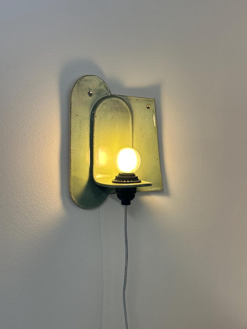 May include: A wall-mounted light fixture featuring a pale green, speckled ceramic backplate with an arched design. A small, round, glowing light bulb sits on a horizontal shelf extending from the center, connected to a black base and a white cord.
