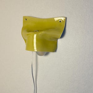 May include: A wall-mounted, yellow ceramic light fixture with a bulb and a silver wire. The fixture has a unique, curved shape with two holes for mounting. The light is a bright, modern design.