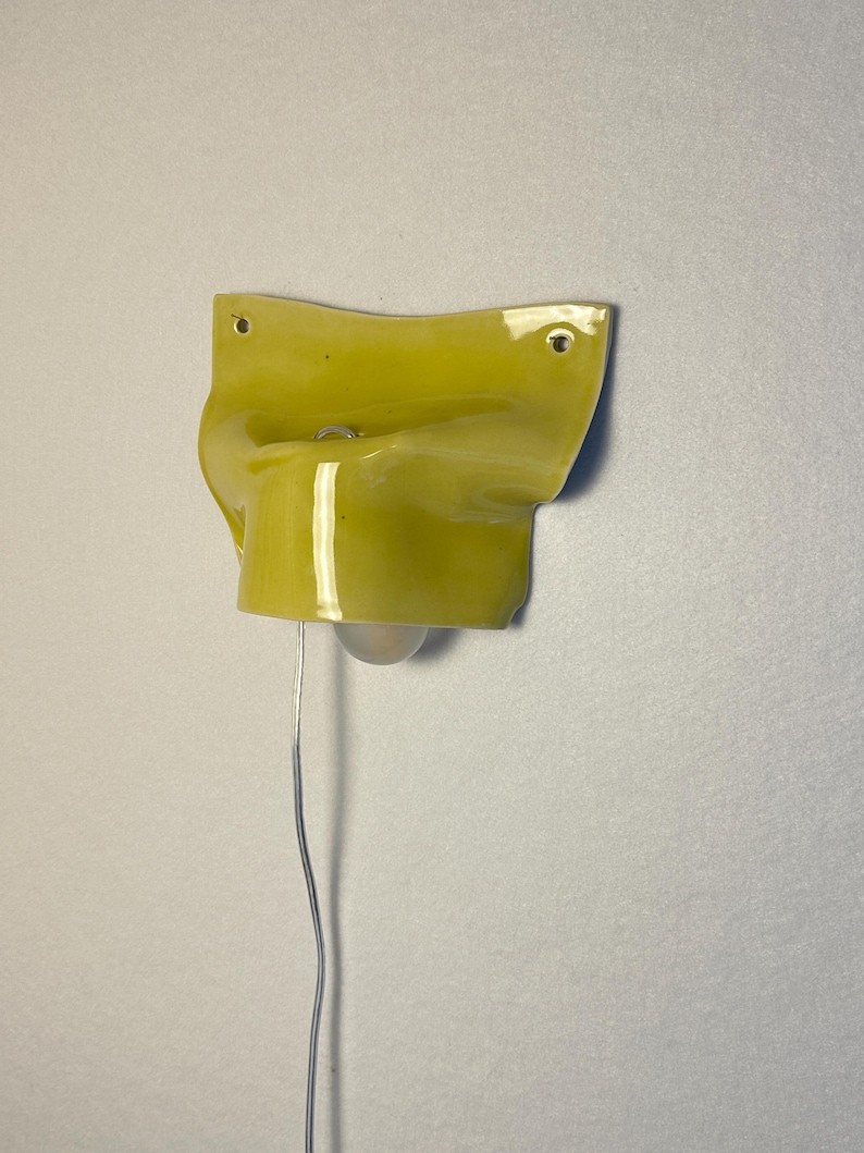 May include: A wall-mounted, yellow ceramic light fixture with a bulb and a silver wire. The fixture has a unique, curved shape with two holes for mounting. The light is a bright, modern design.