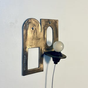 K&ouml;nnte beinhalten: Eine Wandleuchte mit einer flachen, bronzefarbenen Metallplatte, die oben abgerundet ist und einen rechteckigen Ausschnitt aufweist. Eine kleine, runde wei&szlig;e Gl&uuml;hbirne sitzt auf einer horizontalen Ablage an der Platte, verbunden durch ein d&uuml;nnes wei&szlig;es Kabel.