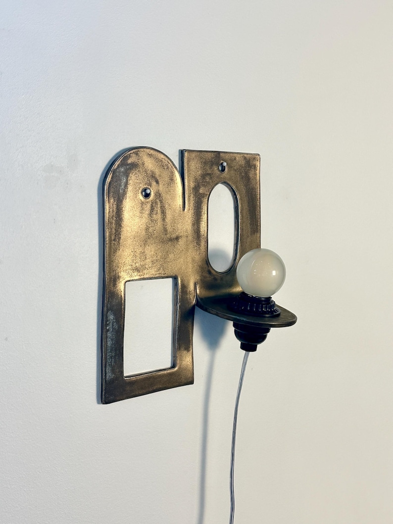 K&ouml;nnte beinhalten: Eine Wandleuchte mit einer flachen, bronzefarbenen Metallplatte, die oben abgerundet ist und einen rechteckigen Ausschnitt aufweist. Eine kleine, runde wei&szlig;e Gl&uuml;hbirne sitzt auf einer horizontalen Ablage an der Platte, verbunden durch ein d&uuml;nnes wei&szlig;es Kabel.