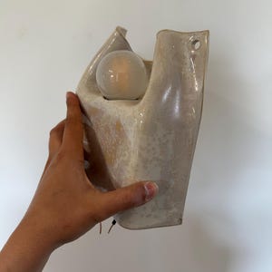 May include: A wall-mounted ceramic sconce with a glossy, speckled cream and beige glaze. The fixture features an organic, sculptural shape with an open top housing a single round, frosted light bulb and a small circular hole for mounting.