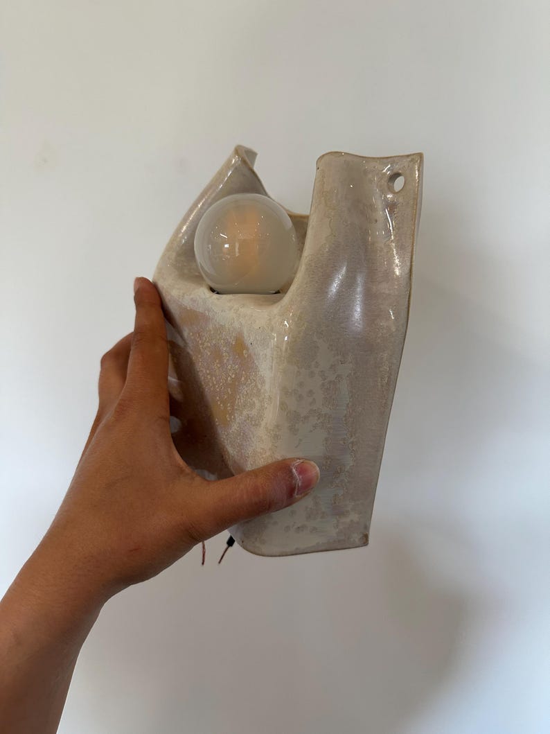 May include: A wall-mounted ceramic sconce with a glossy, speckled cream and beige glaze. The fixture features an organic, sculptural shape with an open top housing a single round, frosted light bulb and a small circular hole for mounting.