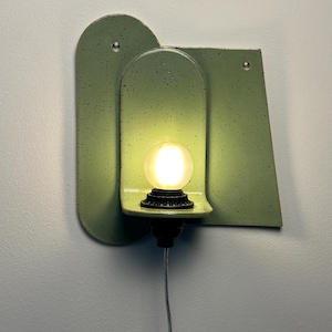 May include: A wall-mounted light fixture featuring a sage green speckled ceramic backplate with an arched cutout. A small black socket holds a glowing, round, frosted light bulb. The fixture includes two small circular holes for mounting purposes.