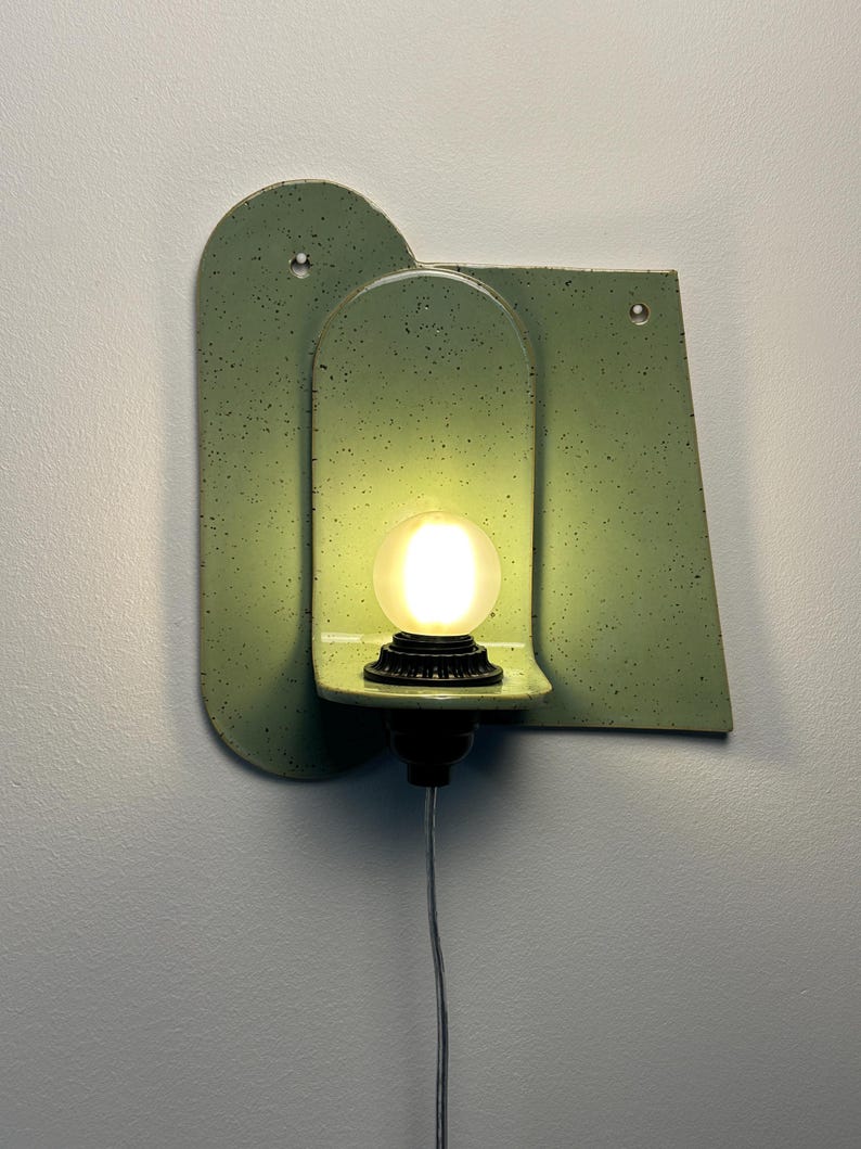 May include: A wall-mounted light fixture featuring a sage green speckled ceramic backplate with an arched cutout. A small black socket holds a glowing, round, frosted light bulb. The fixture includes two small circular holes for mounting purposes.