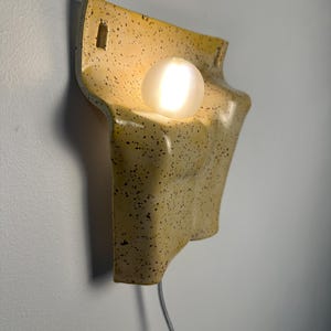 May include: A wall-mounted ceramic lamp with a glossy cream-colored glaze and dark brown speckles. The fixture features a curved, molded shape with two rectangular mounting holes at the top and a single glowing white spherical light bulb.
