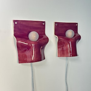 May include: Two wall-mounted sconces featuring a glossy, deep magenta ceramic base with a fluid, draped silhouette. Each fixture includes a central, spherical frosted glass light bulb and a clear power cord extending from the bottom edge.