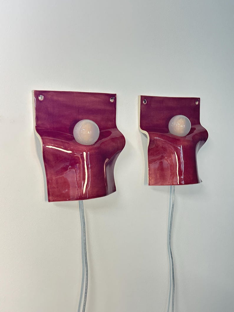 May include: Two wall-mounted sconces featuring a glossy, deep magenta ceramic base with a fluid, draped silhouette. Each fixture includes a central, spherical frosted glass light bulb and a clear power cord extending from the bottom edge.