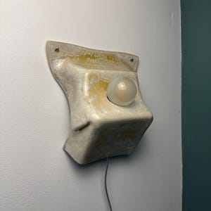 May include: A wall-mounted ceramic light fixture with a glossy, off-white and yellow mottled glaze. The structure features a flared top edge with two mounting holes and a central circular opening holding a single, spherical, frosted glass bulb.