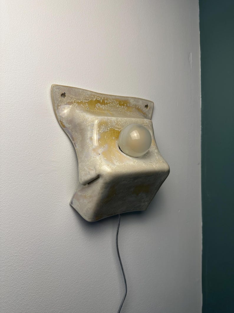 May include: A wall-mounted ceramic light fixture with a glossy, off-white and yellow mottled glaze. The structure features a flared top edge with two mounting holes and a central circular opening holding a single, spherical, frosted glass bulb.