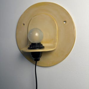 Pu&ograve; includere: Una lampada da parete con una piastra posteriore circolare in ceramica giallo pallido dotata di due piccoli fori. Un portalampada nero sostiene una lampadina rotonda in vetro smerigliato su una piccola mensola in ceramica. Un cavo nero pende.