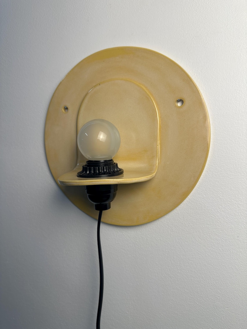 Pu&ograve; includere: Una lampada da parete con una piastra posteriore circolare in ceramica giallo pallido dotata di due piccoli fori. Un portalampada nero sostiene una lampadina rotonda in vetro smerigliato su una piccola mensola in ceramica. Un cavo nero pende.