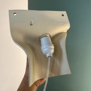 Pode incluir: Um candeeiro de parede de cor creme com um abajur de pl&aacute;stico moldado e curvo, apresentando dois orif&iacute;cios de montagem circulares no topo. Um casquilho de l&acirc;mpada branco est&aacute; centrado no meio, ligado a um cabo el&eacute;trico transparente.
