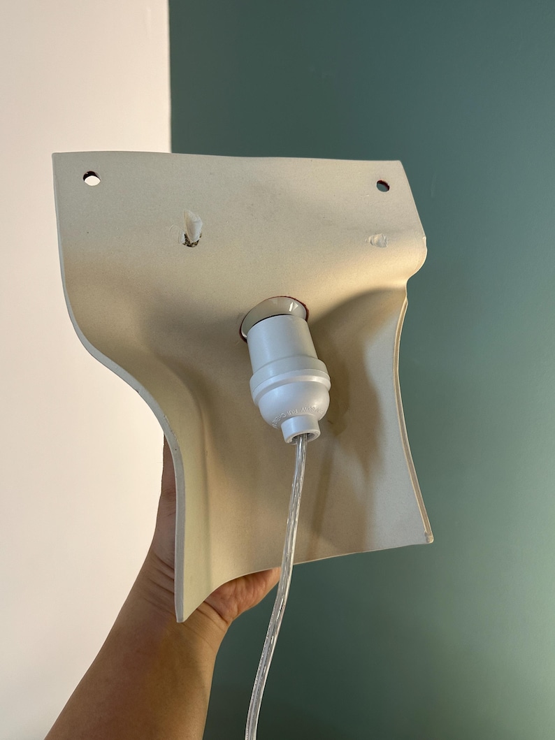 Pode incluir: Um candeeiro de parede de cor creme com um abajur de pl&aacute;stico moldado e curvo, apresentando dois orif&iacute;cios de montagem circulares no topo. Um casquilho de l&acirc;mpada branco est&aacute; centrado no meio, ligado a um cabo el&eacute;trico transparente.