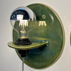 May include: A wall-mounted light fixture featuring a circular, glossy green ceramic backplate with a small integrated shelf. A black socket holds a clear glass light bulb with a reflective silver top half and visible internal filaments.
