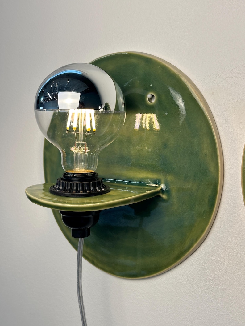 May include: A wall-mounted light fixture featuring a circular, glossy green ceramic backplate with a small integrated shelf. A black socket holds a clear glass light bulb with a reflective silver top half and visible internal filaments.
