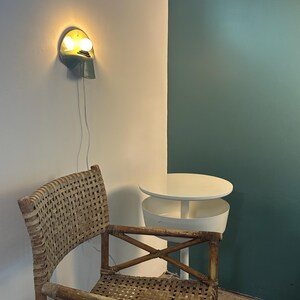 Puede incluir: Una l&aacute;mpara de pared con acabado met&aacute;lico y dos bombillas encendidas, situada sobre una silla de rat&aacute;n trenzado con estructura de madera. Junto a la silla hay una mesa auxiliar redonda blanca con un dise&ntilde;o de niveles y una base central.