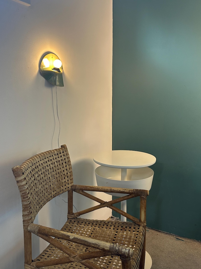 Puede incluir: Una l&aacute;mpara de pared con acabado met&aacute;lico y dos bombillas encendidas, situada sobre una silla de rat&aacute;n trenzado con estructura de madera. Junto a la silla hay una mesa auxiliar redonda blanca con un dise&ntilde;o de niveles y una base central.