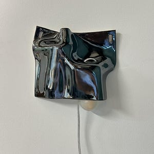 May include: A wall-mounted lamp featuring a dark, reflective, metallic-finished shade with an irregular, rippled, and fluid surface texture. The lamp includes a single exposed light bulb extending from the bottom and a thin, light-colored power cord.
