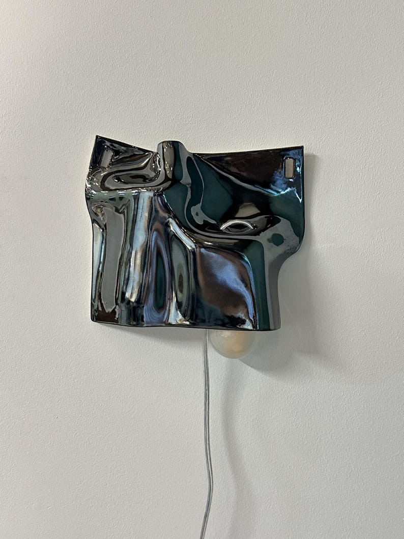 May include: A wall-mounted lamp featuring a dark, reflective, metallic-finished shade with an irregular, rippled, and fluid surface texture. The lamp includes a single exposed light bulb extending from the bottom and a thin, light-colored power cord.