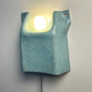 May include: A light blue ceramic wall sconce with a speckled finish and a smooth, glossy texture. The fixture features a curved, folded design with two circular mounting holes at the top corners and a single glowing round light bulb centered.