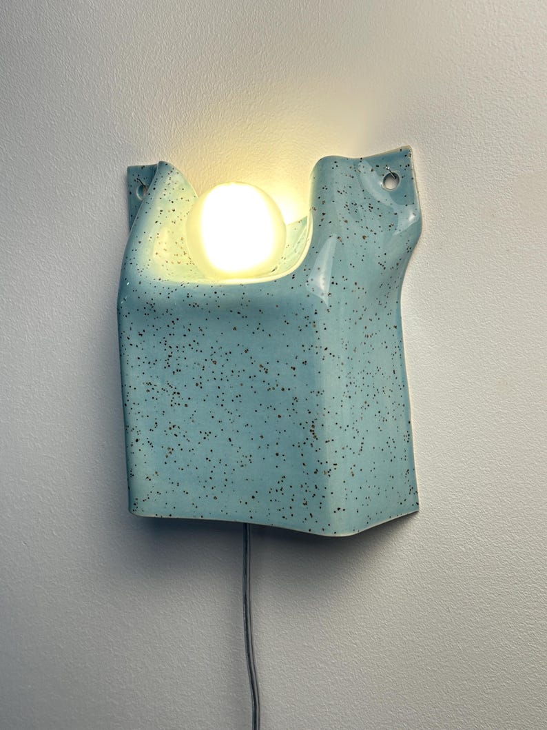 May include: A light blue ceramic wall sconce with a speckled finish and a smooth, glossy texture. The fixture features a curved, folded design with two circular mounting holes at the top corners and a single glowing round light bulb centered.