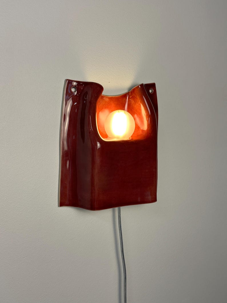 Puede incluir: Un aplique de pared de cer&aacute;mica de color rojo intenso con acabado brillante y forma org&aacute;nica curva. La pieza presenta un recorte central que alberga una bombilla esf&eacute;rica brillante. Dos peque&ntilde;os ojales met&aacute;licos son visibles en la parte superior.