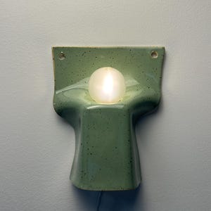 May include: A wall-mounted ceramic light fitting in a speckled sage green glaze. The piece features a curved, moulded form with a central circular cutout holding a glowing white spherical light bulb. Two small holes are visible at the top corners.