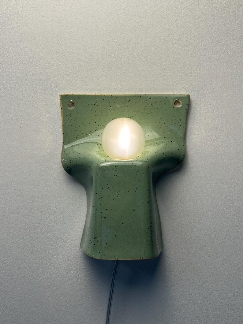 May include: A wall-mounted ceramic light fitting in a speckled sage green glaze. The piece features a curved, moulded form with a central circular cutout holding a glowing white spherical light bulb. Two small holes are visible at the top corners.