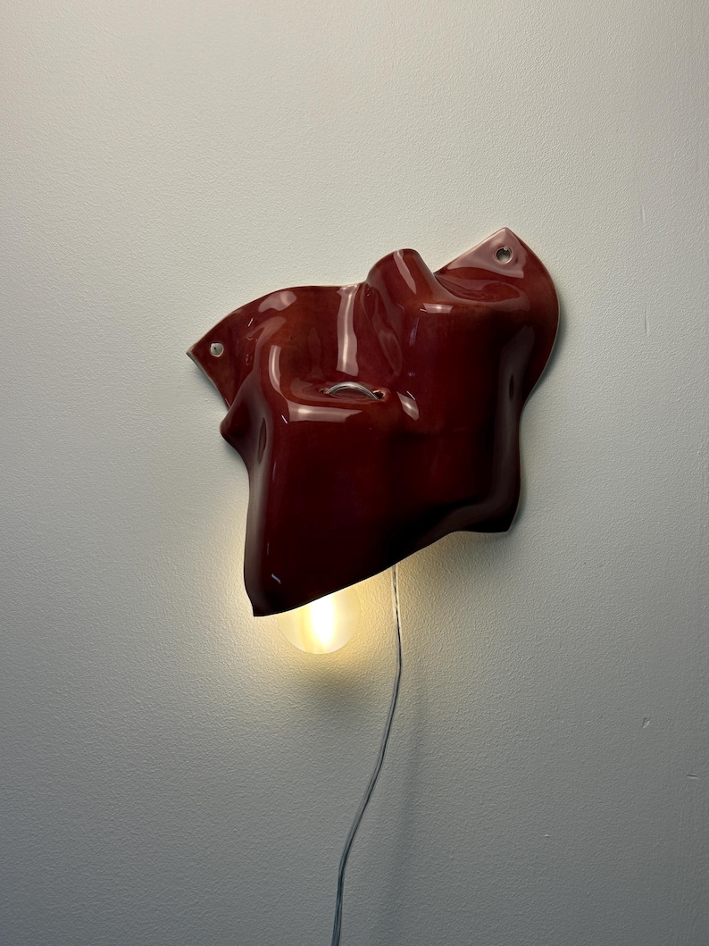 Puede incluir: Una l&aacute;mpara de pared con una forma org&aacute;nica de cer&aacute;mica o resina en color rojo oscuro brillante y contornos irregulares. Una bombilla de luz c&aacute;lida sobresale del borde inferior, conectada por un cable el&eacute;ctrico fino y claro.