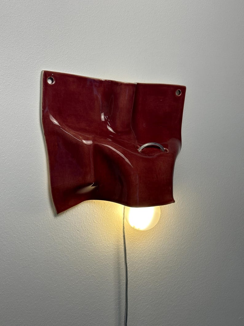 May include: A wall-mounted ceramic light fixture with a glossy deep red finish and an irregular, undulating surface. The piece features two small circular holes for mounting and a silver metal ring detail. A single glowing light bulb hangs below.
