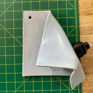 May include: A light gray ceramic wall sconce with a folded, triangular silhouette and a single circular mounting hole. The fixture includes a black socket for a light bulb, positioned on a green cutting mat with imperial measurement markings.