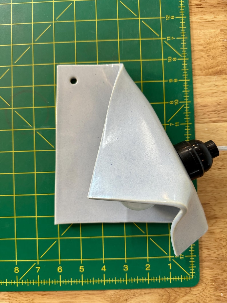 May include: A light gray ceramic wall sconce with a folded, triangular silhouette and a single circular mounting hole. The fixture includes a black socket for a light bulb, positioned on a green cutting mat with imperial measurement markings.