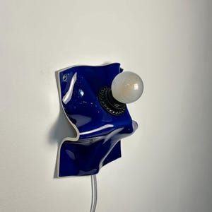 May include: A wall-mounted lamp featuring a glossy, deep blue ceramic base with an irregular, wavy, and folded shape. A black circular socket is centered on the ceramic, holding a single, round, frosted white light bulb. A thin, clear power cord hangs down.
