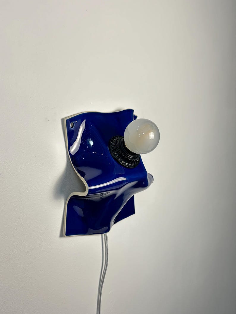 May include: A wall-mounted lamp featuring a glossy, deep blue ceramic base with an irregular, wavy, and folded shape. A black circular socket is centered on the ceramic, holding a single, round, frosted white light bulb. A thin, clear power cord hangs down.