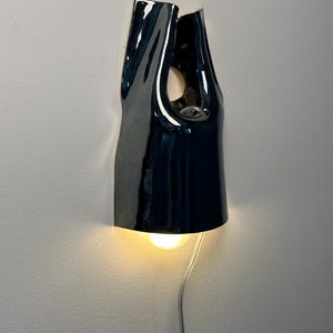 May include: A glossy black ceramic wall sconce with an organic, folded silhouette. The fixture features a central cutout and a downward-facing light bulb that emits a warm glow. A thin, light-coloured electrical cord hangs from the bottom of the lamp.