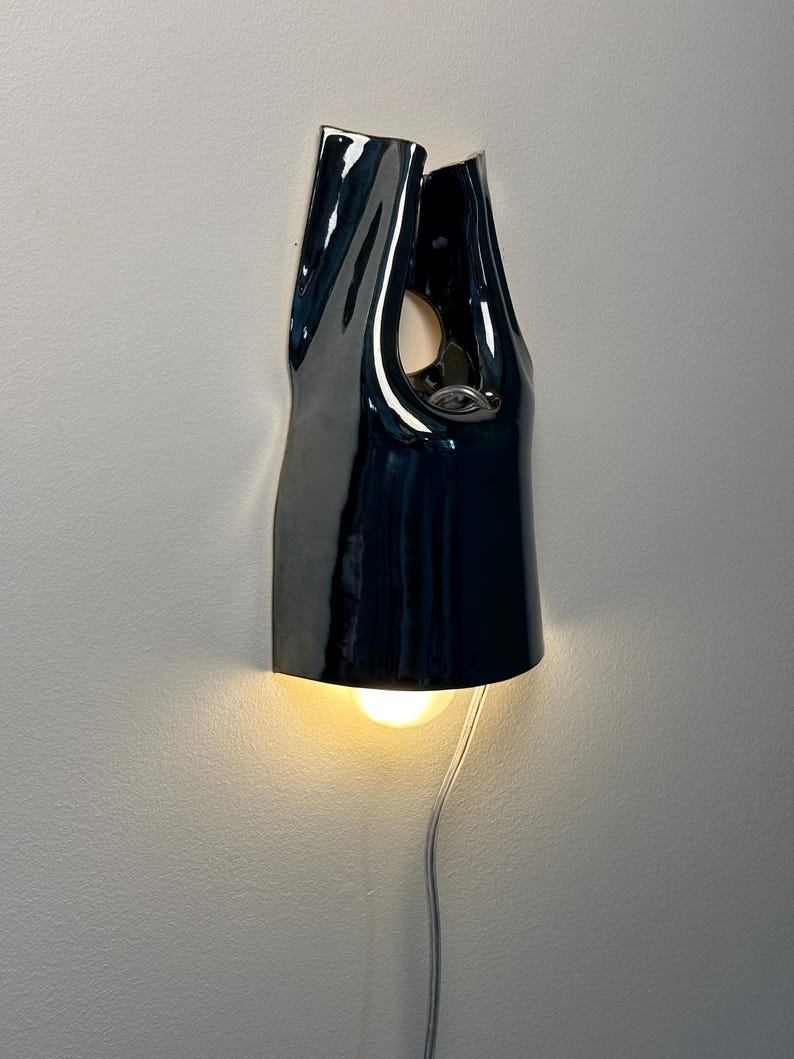 May include: A glossy black ceramic wall sconce with an organic, folded silhouette. The fixture features a central cutout and a downward-facing light bulb that emits a warm glow. A thin, light-coloured electrical cord hangs from the bottom of the lamp.