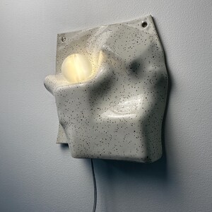 May include: A wall-mounted ceramic lamp with an off-white, speckled finish and an irregular, molded surface. The fixture features a circular, glowing light bulb nestled within a recessed cavity on the left side and a black power cord extending downward.