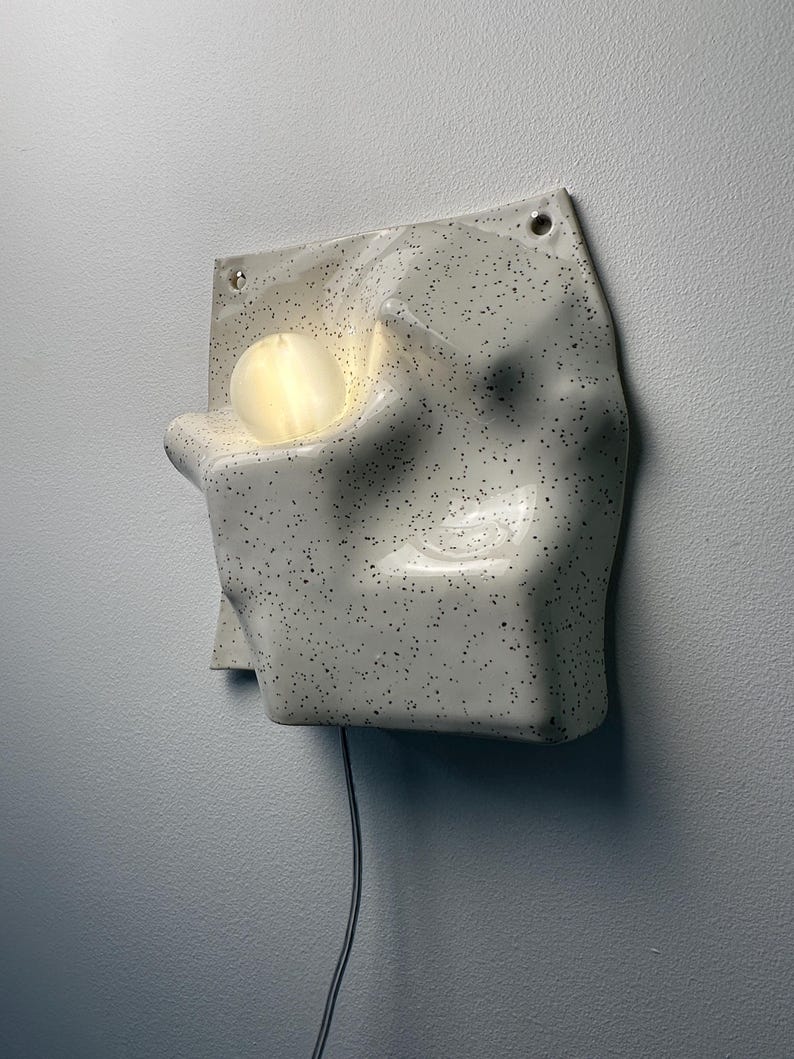 May include: A wall-mounted ceramic lamp with an off-white, speckled finish and an irregular, molded surface. The fixture features a circular, glowing light bulb nestled within a recessed cavity on the left side and a black power cord extending downward.