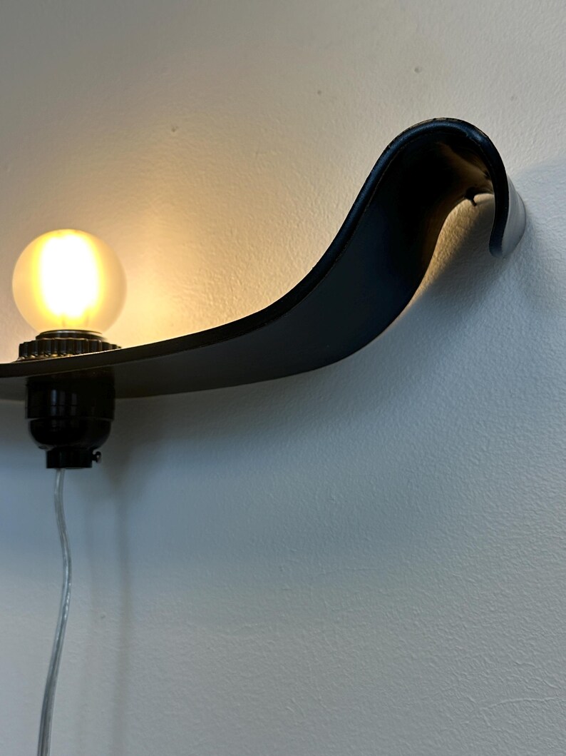 May include: A black, wall-mounted light fixture with a curved, wave-like design. The fixture holds a round, illuminated light bulb. The light is attached to the wall with a black base and a silver cord.