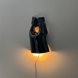 May include: A glossy black ceramic wall light with a sculptural, elongated form featuring a central cutout. The fixture emits a warm, glowing light from its base, illuminating the wall below, and includes a thin white power cord extending downwards.