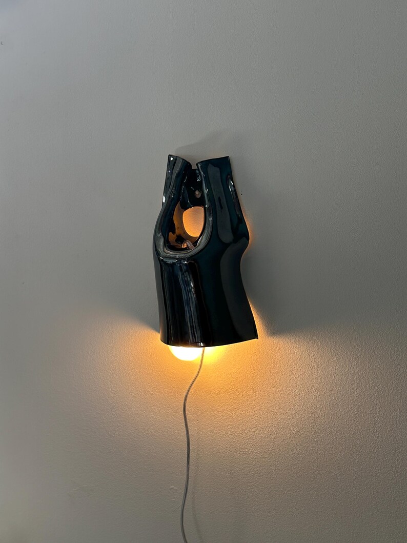 May include: A glossy black ceramic wall light with a sculptural, elongated form featuring a central cutout. The fixture emits a warm, glowing light from its base, illuminating the wall below, and includes a thin white power cord extending downwards.