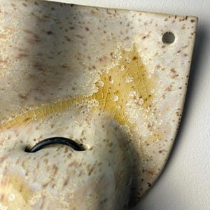 May include: A curved ceramic wall piece featuring a cream-colored glaze with brown speckles and a yellow crystalline crackle pattern. The object includes a small circular hole in the upper corner and a black loop attached to the center surface.