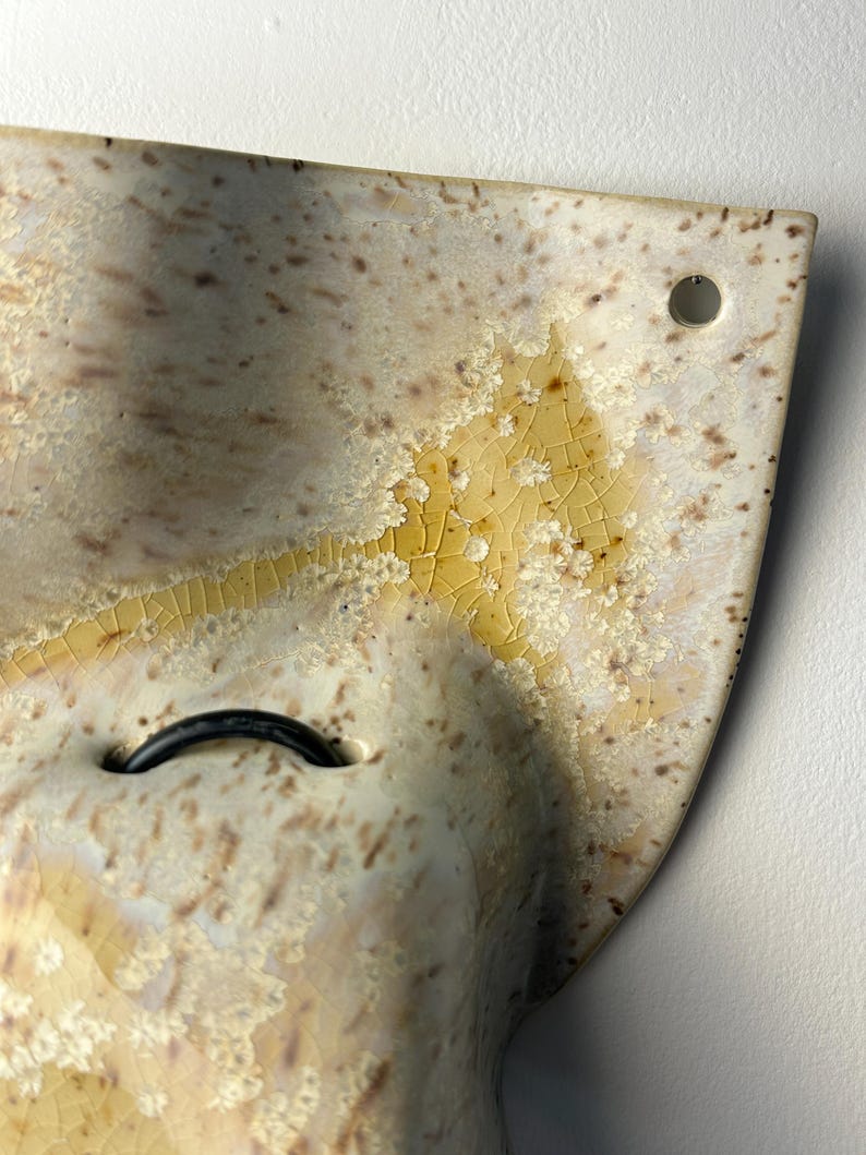 May include: A curved ceramic wall piece featuring a cream-colored glaze with brown speckles and a yellow crystalline crackle pattern. The object includes a small circular hole in the upper corner and a black loop attached to the center surface.
