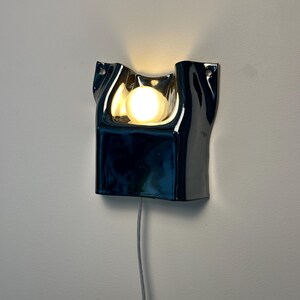 May include: A wall-mounted lamp featuring a glossy, dark blue, undulating ceramic frame with a central cutout. A single round, warm-glowing light bulb is positioned within the recessed area. A thin, light-colored power cord hangs from the bottom.