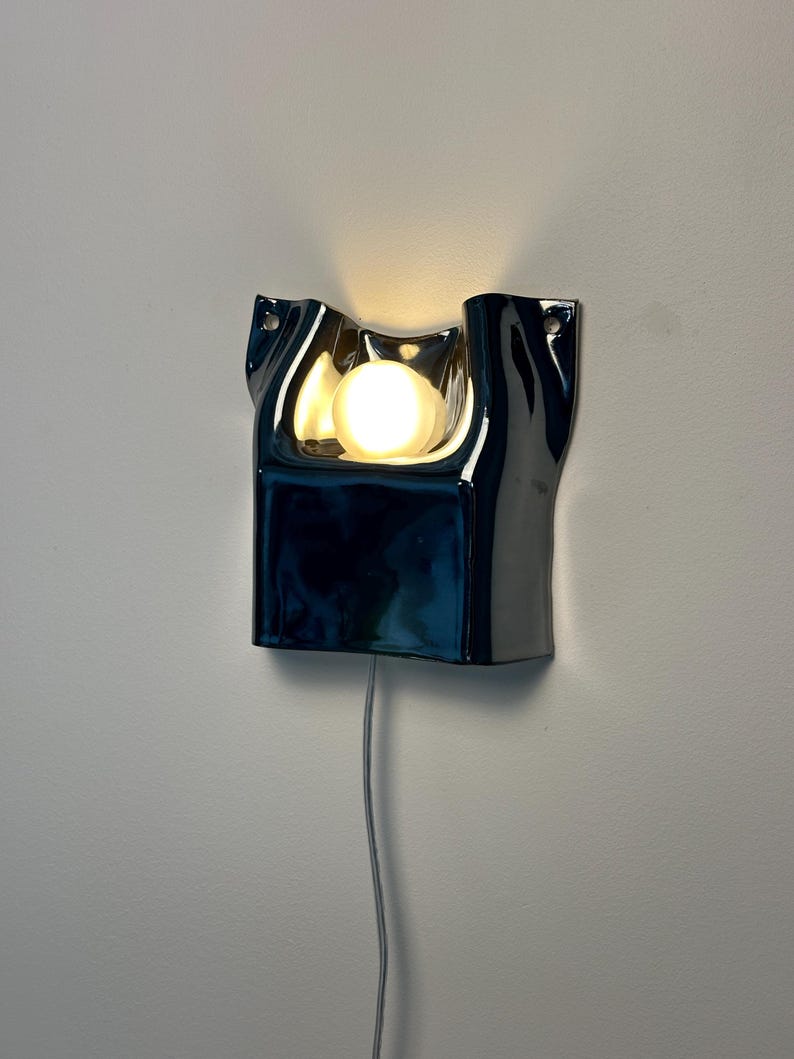 May include: A wall-mounted lamp featuring a glossy, dark blue, undulating ceramic frame with a central cutout. A single round, warm-glowing light bulb is positioned within the recessed area. A thin, light-colored power cord hangs from the bottom.