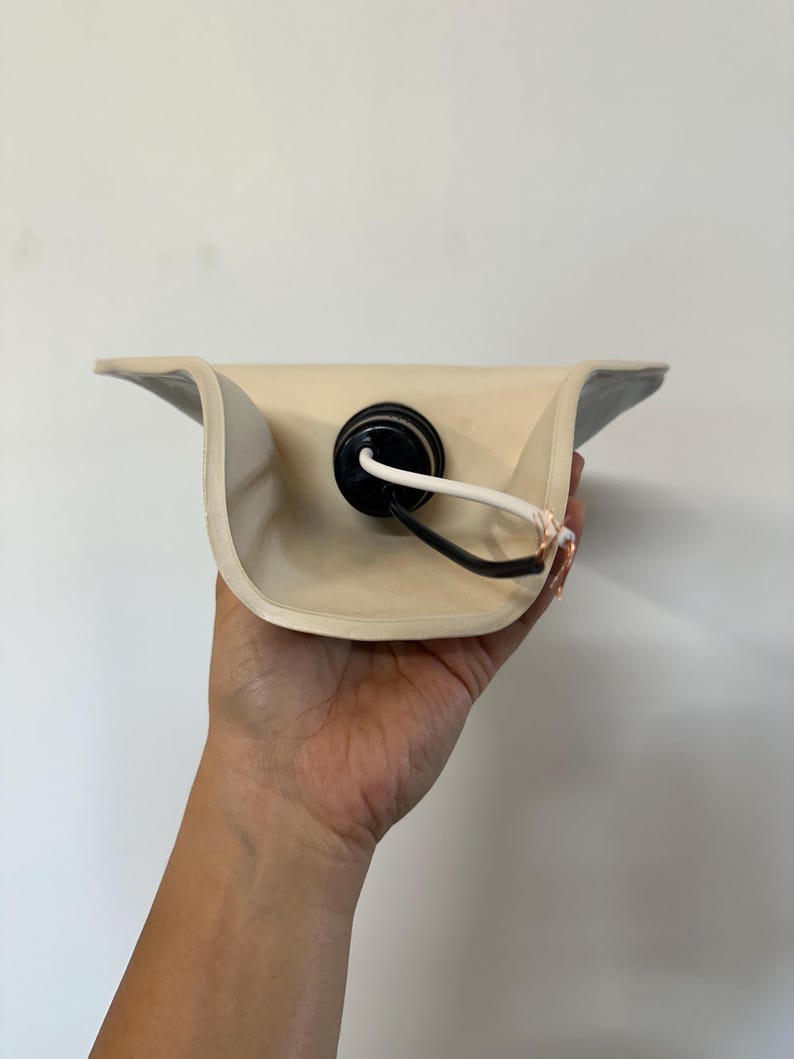 May include: A cream-colored curved ceramic lamp shade featuring a central black socket housing. A white electrical cord with exposed copper wiring extends from the socket, which is mounted in the center of the concave interior of the shade.