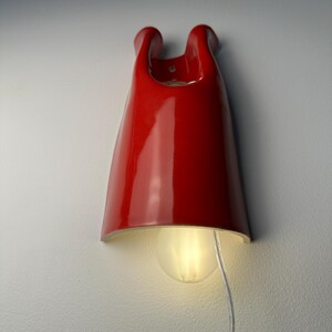 May include: A glossy red ceramic wall sconce with a curved, elongated silhouette. The fixture features a central opening at the top and a rounded light bulb emitting a soft glow from the bottom. A thin, clear electrical cord hangs downward.