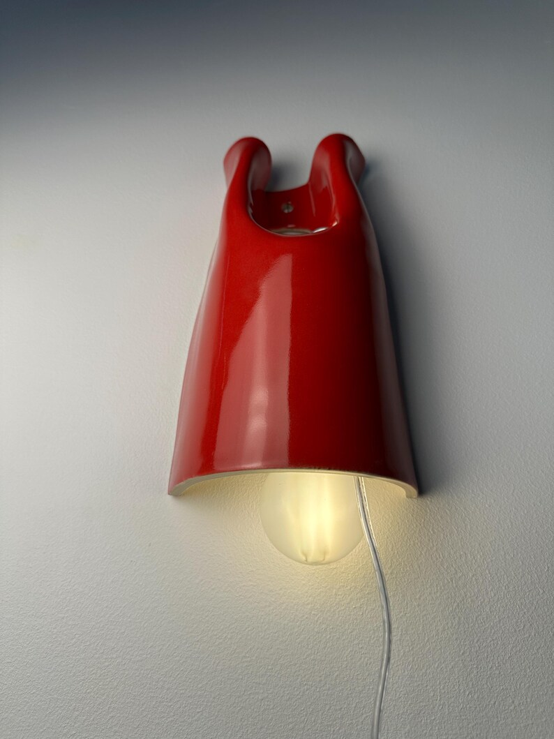 May include: A glossy red ceramic wall sconce with a curved, elongated silhouette. The fixture features a central opening at the top and a rounded light bulb emitting a soft glow from the bottom. A thin, clear electrical cord hangs downward.
