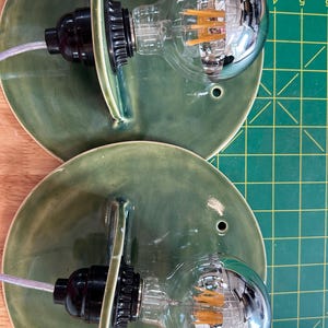 May include: Two wall sconces featuring circular, moss-green glazed ceramic backplates with a single mounting hole. Each fixture includes a black socket holding a large, clear globe-shaped LED filament light bulb with a silver-tinted top section.